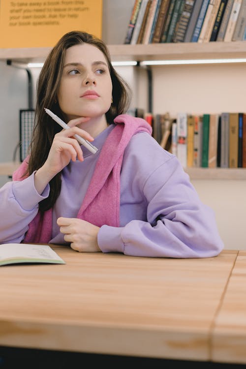 Clever young female student with long dark hair in trendy clothes touching chin with finger and looking away while thinking about task during lesson in university