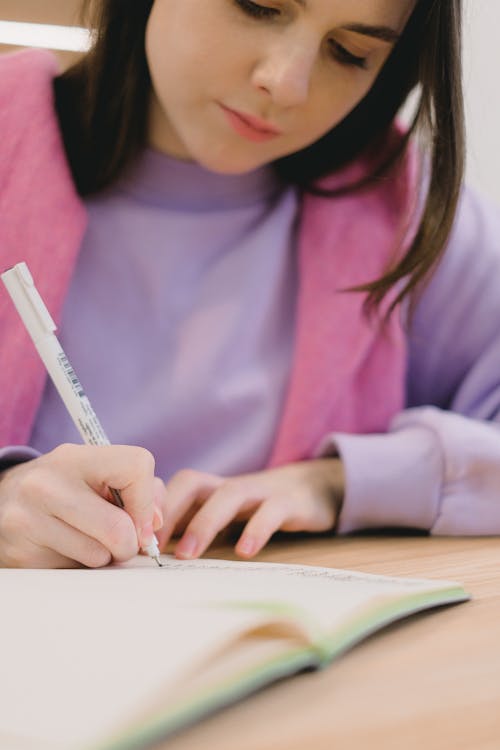 Free Crop young female student with long dark hair in casual outfit taking notes in copybook while preparing for exam sitting at wooden table in classroom Stock Photo