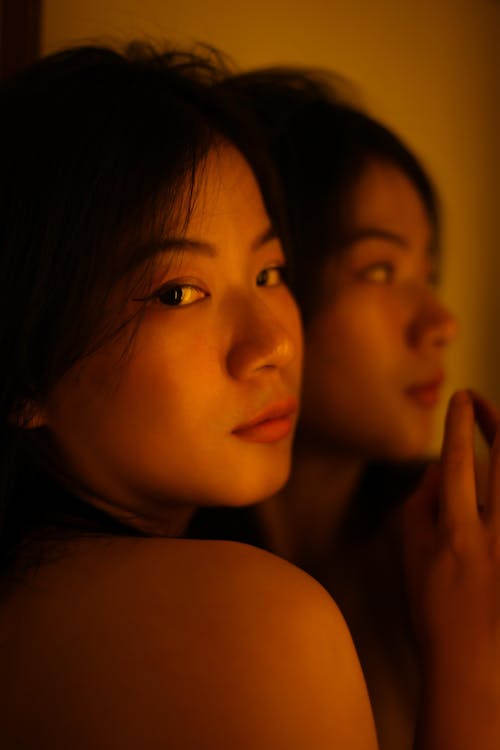 Free Tender alluring ethnic female looking at camera in dim yellow light of dark studio with mirror Stock Photo