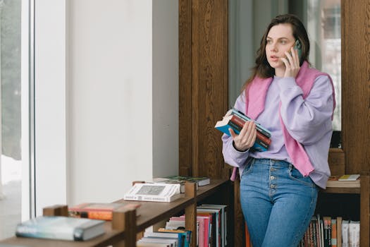 Woman standing in a library, holding books and talking on the phone indoors.