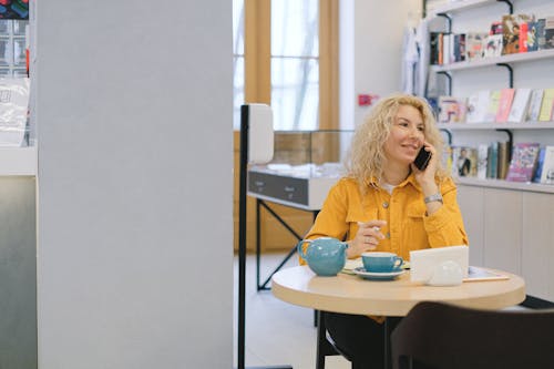 Free Woman sitting at table in library and drinking tea during phone conversation and looking away Stock Photo