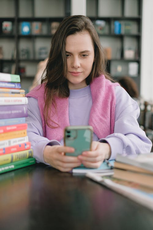 Young female student sitting at table in library and browsing smartphone while preparing for exam