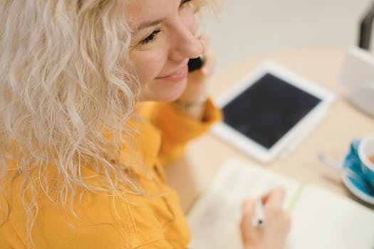 Smiling blonde woman multitasking with a phone and tablet at a cozy indoor workplace.
