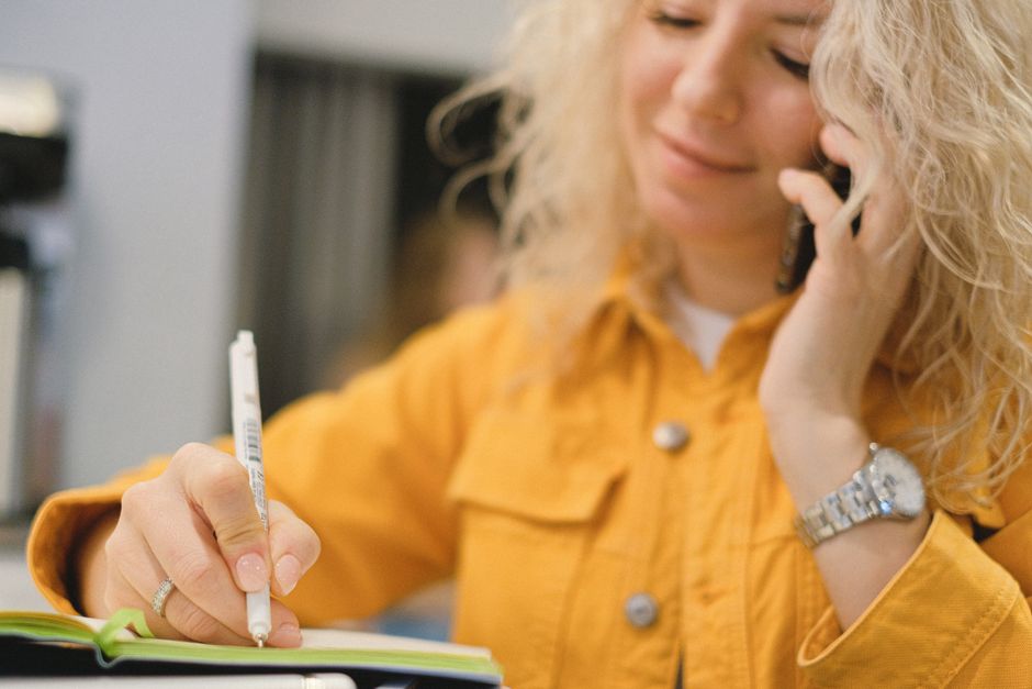 Content blond female employee writing notes while talking on cellphone on blurred background