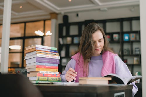 Free Female student in casual clothes sitting at table with books and writing notes while preparing for exam in university library Stock Photo
