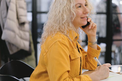 Side view of positive businesswoman sitting at table and writing notes while having conversation on mobile phone