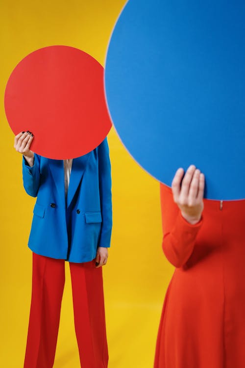 Unrecognizable female models in fashionable outfits covering faces with colored round sheets while standing against yellow background