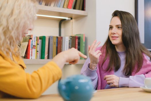 Two women communicate in a library setting, expressing ideas through gestures.
