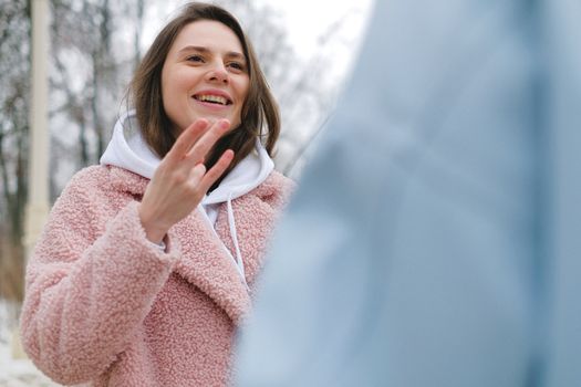 Cheerful female in warm coat looking at partner and showing gesture of sign language while standing in winter park