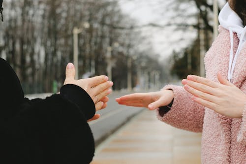 Crop anonymous people in outerwear standing in park on blurred background in winter and using sign language