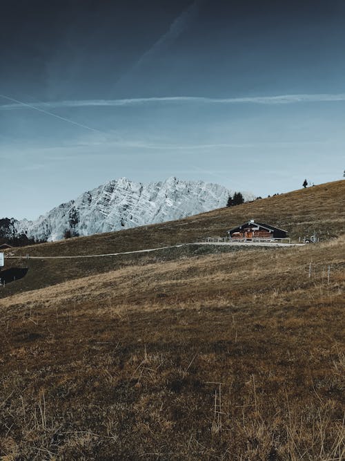 Vast field with rocky mountains and aged timber house under bright blue sky