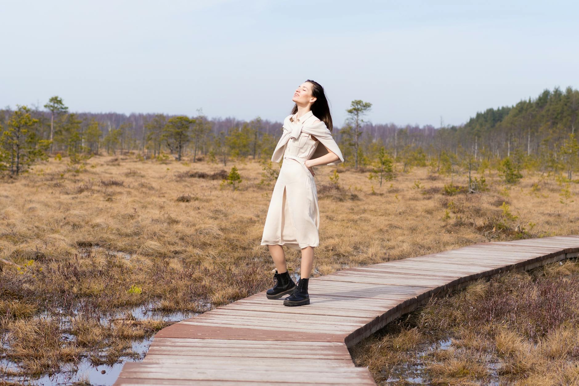 Full body of happy female with eyes closed on wooden path among grass and water in forest