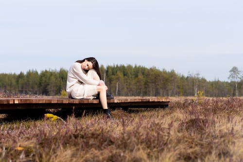 Calm female in casual white dress and black boots relaxing on timber path near dry grass and green forest