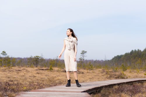 Full body of young slender female with dark hair in trendy white casual dress looking away on timber path