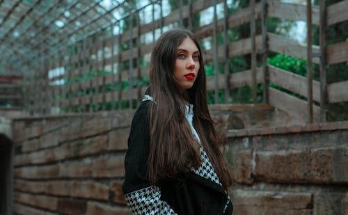 Side view of young female with long straight hair and red lips standing near stone wall and looking away