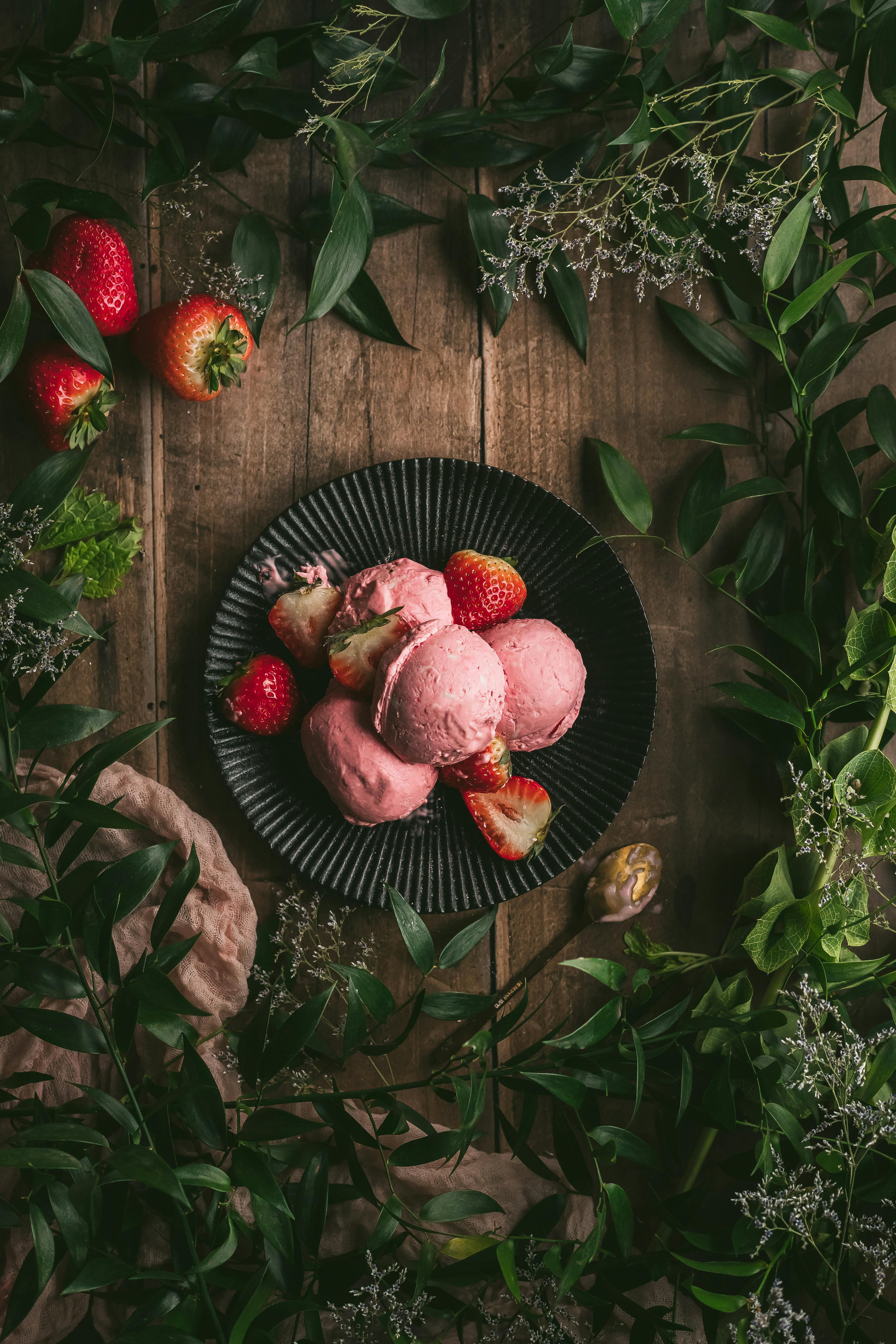 Delicious strawberry ice cream scoops on a black plate surrounded by fresh strawberries and greenery.