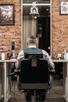 Adult man seen from behind sitting in a barbershop, facing a mirror and brick wall.
