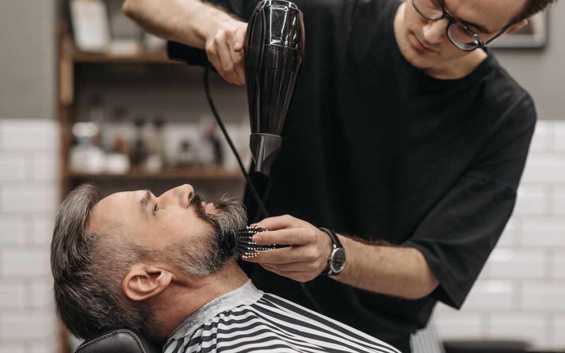 Barber styling a man's beard with hair dryer in a modern salon, capturing the art of grooming.