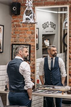 Stylish middle-aged man observing reflection in barber shop mirror.