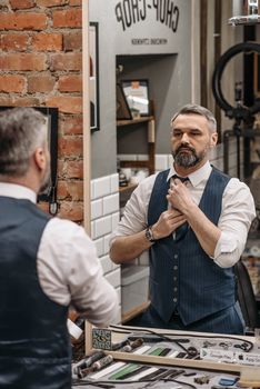 Stylish adult man adjusts his tie while looking in a barbershop mirror, showcasing modern fashion.
