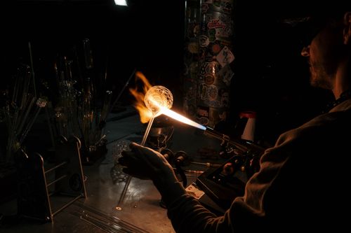 Free An artisan crafts glass using a blowpipe and flame in a dark workshop setting. Stock Photo