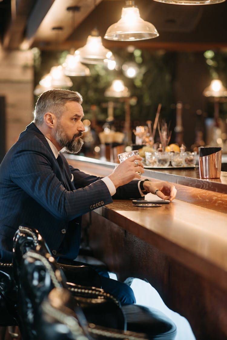 Man In A Suit Holding A Glass Cup While Sitting At The Bar Counter