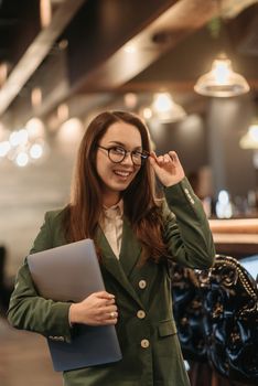 Young woman in glasses holding a laptop, smiling confidently, dressed in elegant green blazer indoors.