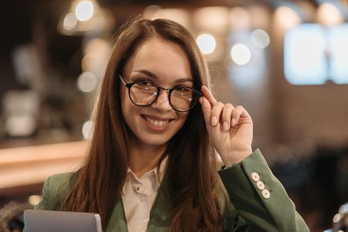 Free Caucasian woman smiling while adjusting her glasses indoors, dressed elegantly in a green jacket. Stock Photo