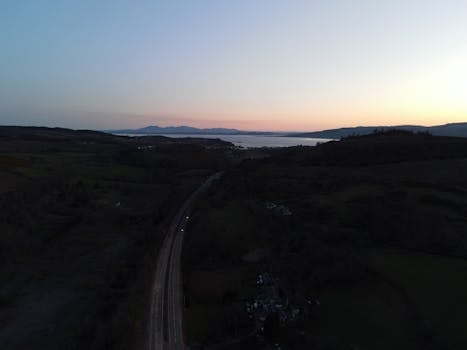 A serene aerial view of the Scottish countryside with a road and sea at sunset.