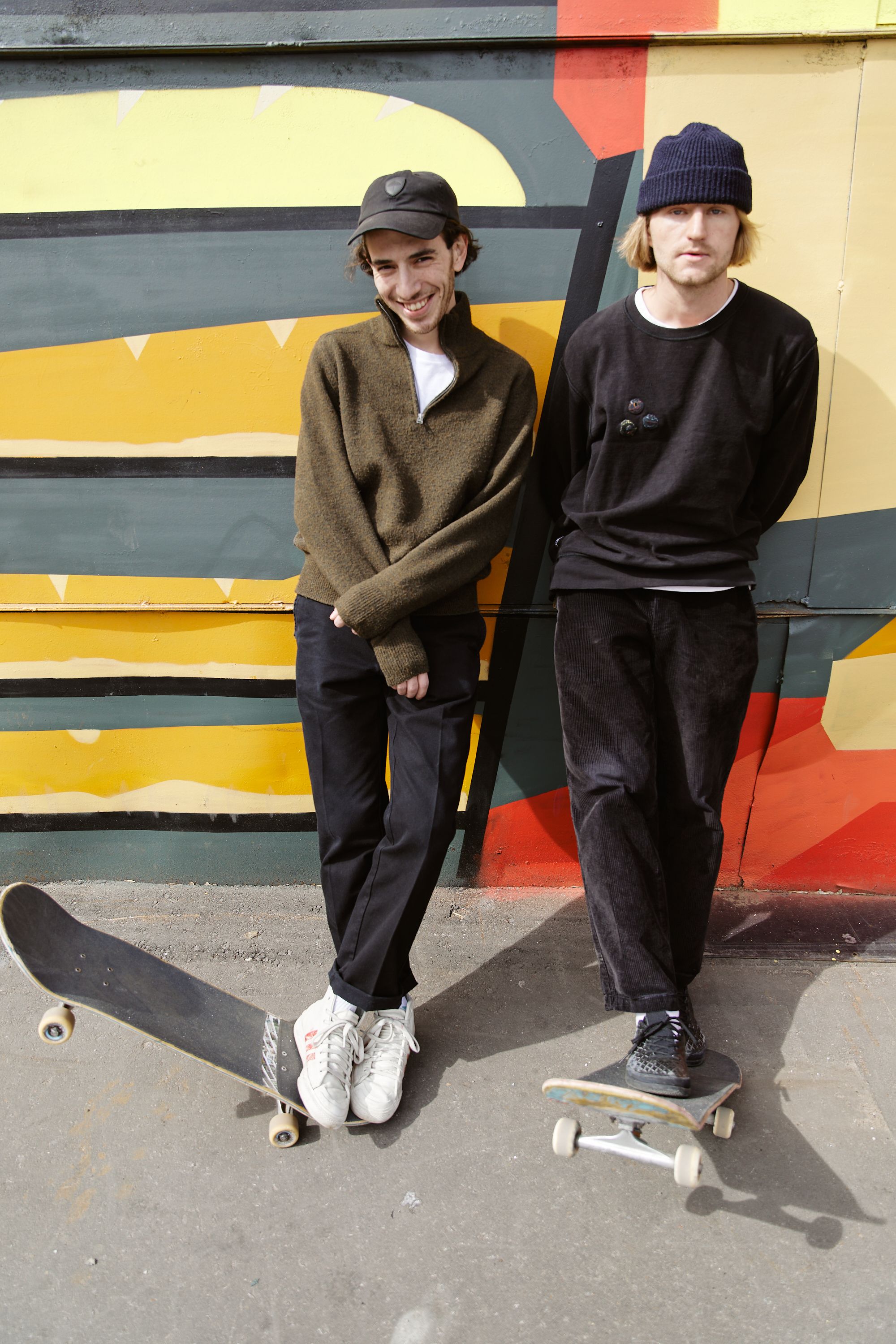 Two skateboarders enjoy a sunny day in front of a colorful street art mural, exuding youthful urban style.