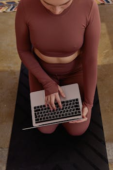 Woman wearing activewear works on a laptop while kneeling on a yoga mat indoors.
