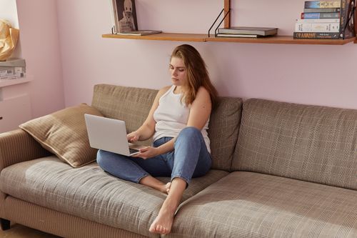 Free Woman in casual attire using a laptop on a sofa, embodying a relaxing work-from-home setup. Stock Photo