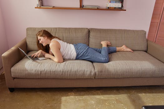 Woman lying on couch, using laptop for remote work, in a cozy home setting.