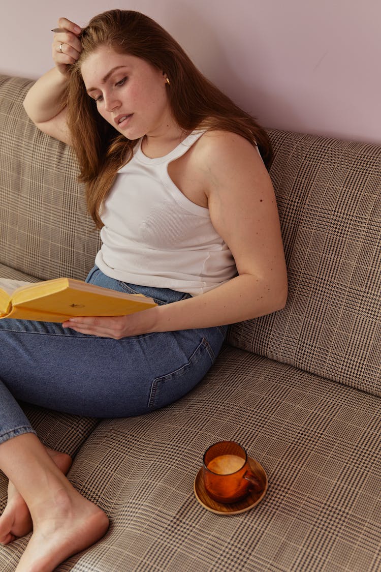 A Woman In White Tank Top Sitting On The Couch While Reading A Book