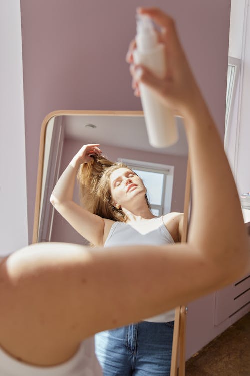 Free A Woman in White Tank Top facing a Mirror Spraying Cosmetic Product on Face Stock Photo