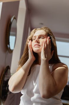 Woman in white tank top applies skincare products while enjoying the sunlight in her home.