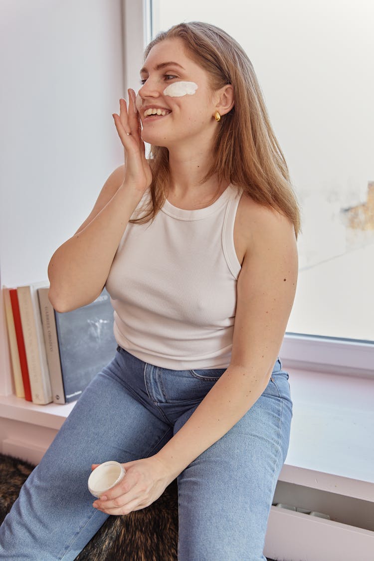 Woman In White Tank Top Putting Face Cream On Her Cheeks 