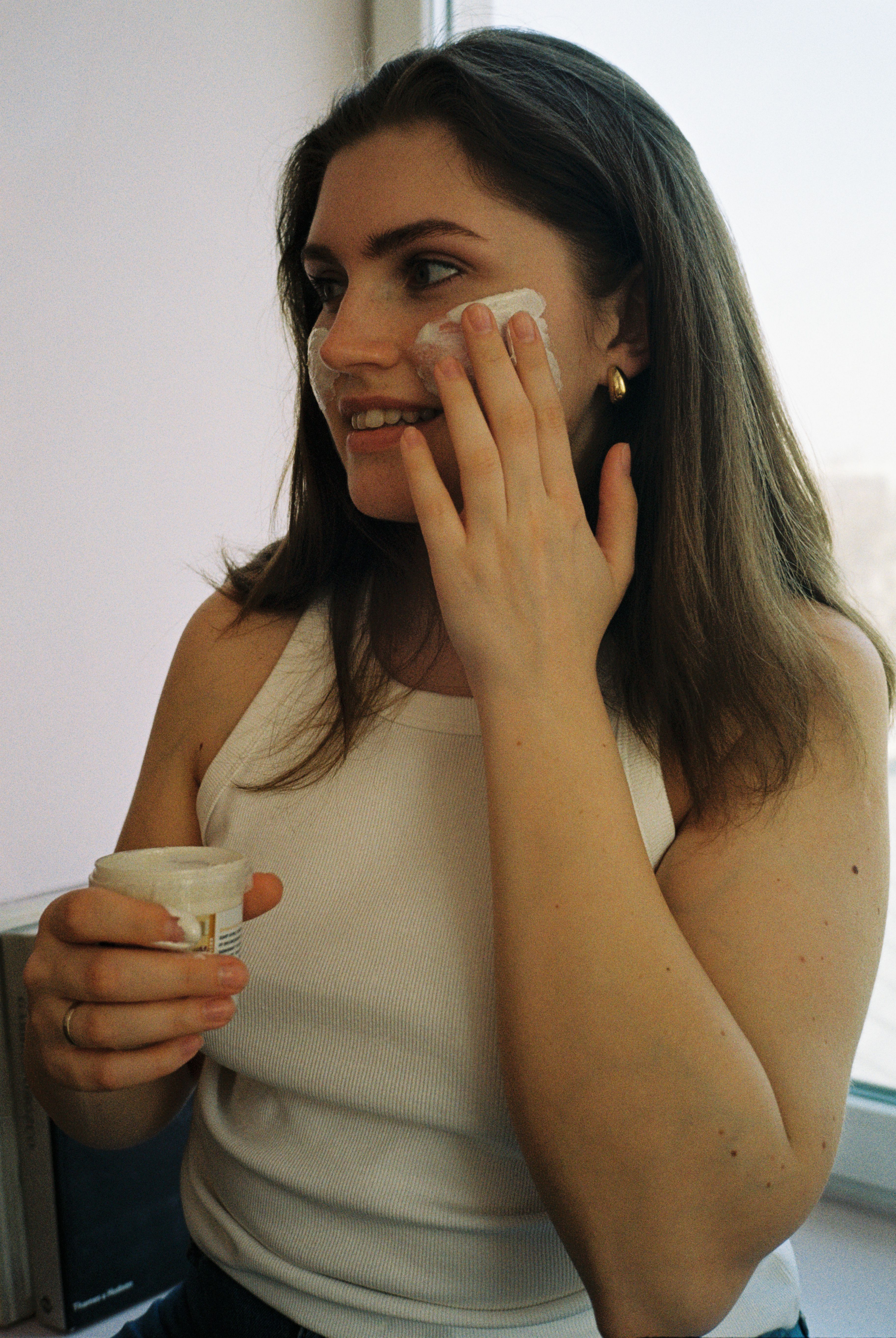 Young woman applying facial cream indoors, promoting skincare routine.