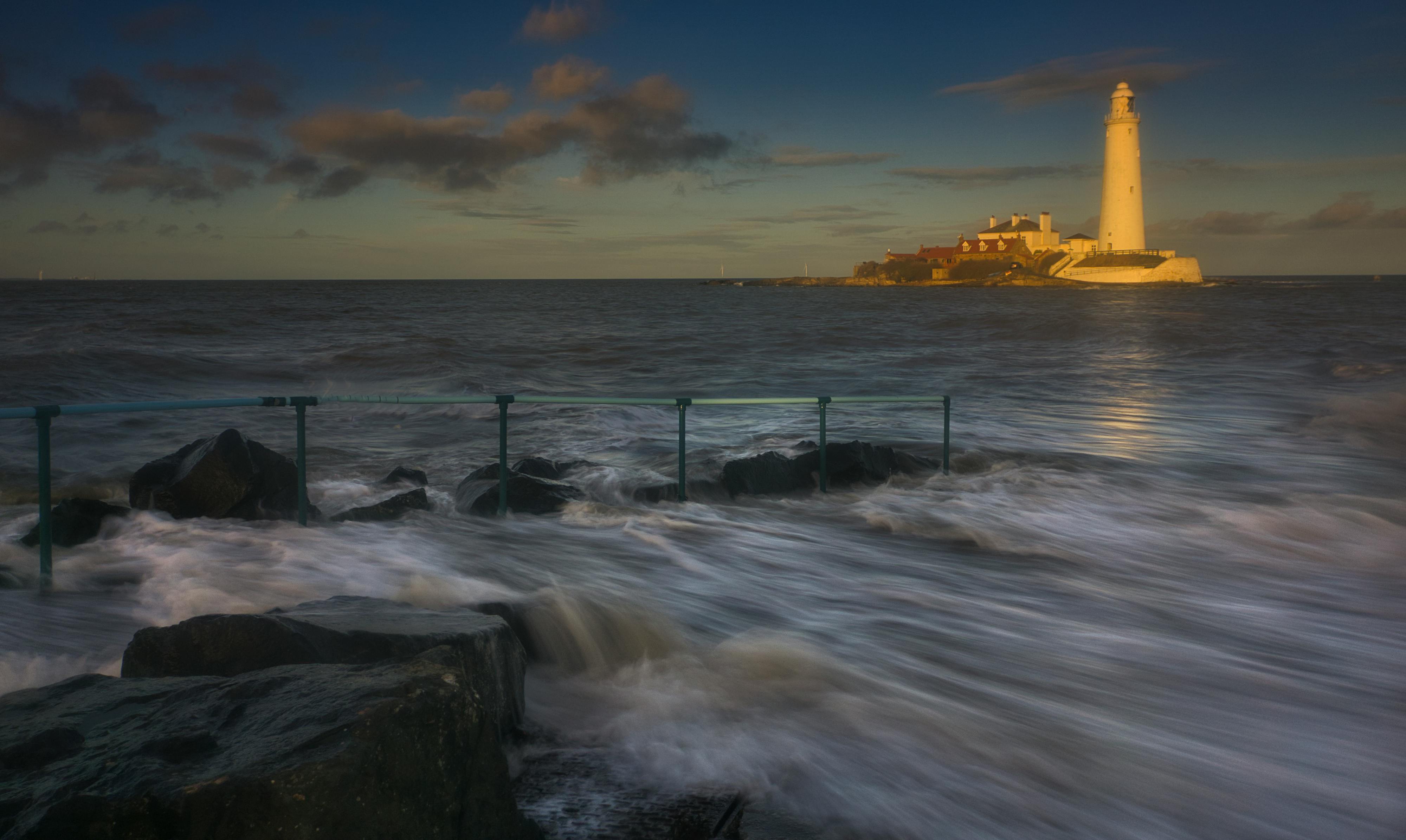 Breathtaking view of waves crashing against rocks with a lighthouse in the background at twilight.