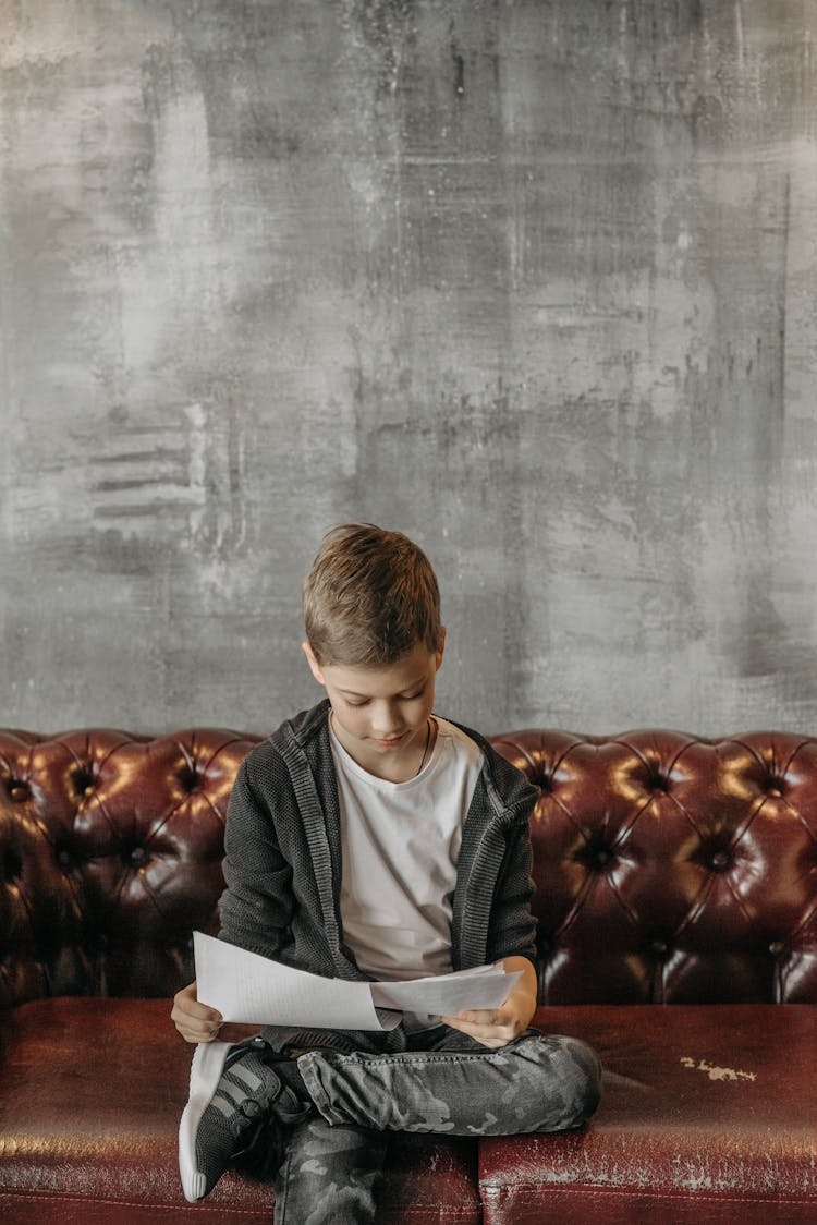 Boy In Gray Jacket Sitting On Brown Couch While Holding White Papers