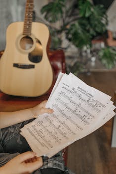 Close-up of hands holding music sheets with an acoustic guitar in the background.