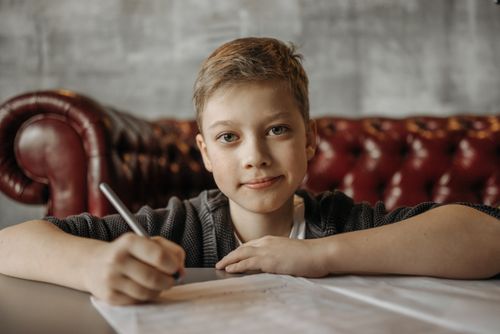 Free Smiling young boy writing on paper seated comfortably on a leather sofa in a cozy indoor setting. Stock Photo