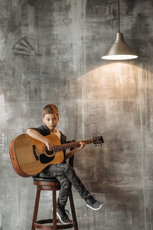 A Young Boy Sitting on a Wooden Chair while Playing Guitar
