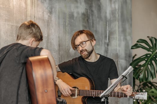 A young boy learning guitar from a bearded man indoors, emphasizing musical guidance.