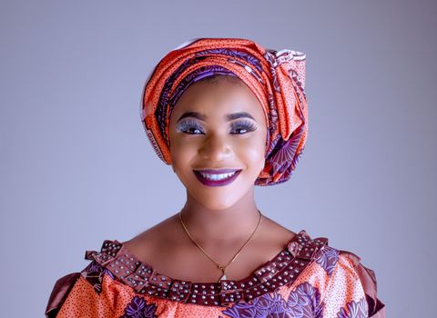 Smiling woman in traditional Nigerian attire with vibrant makeup and a turban, studio portrait.