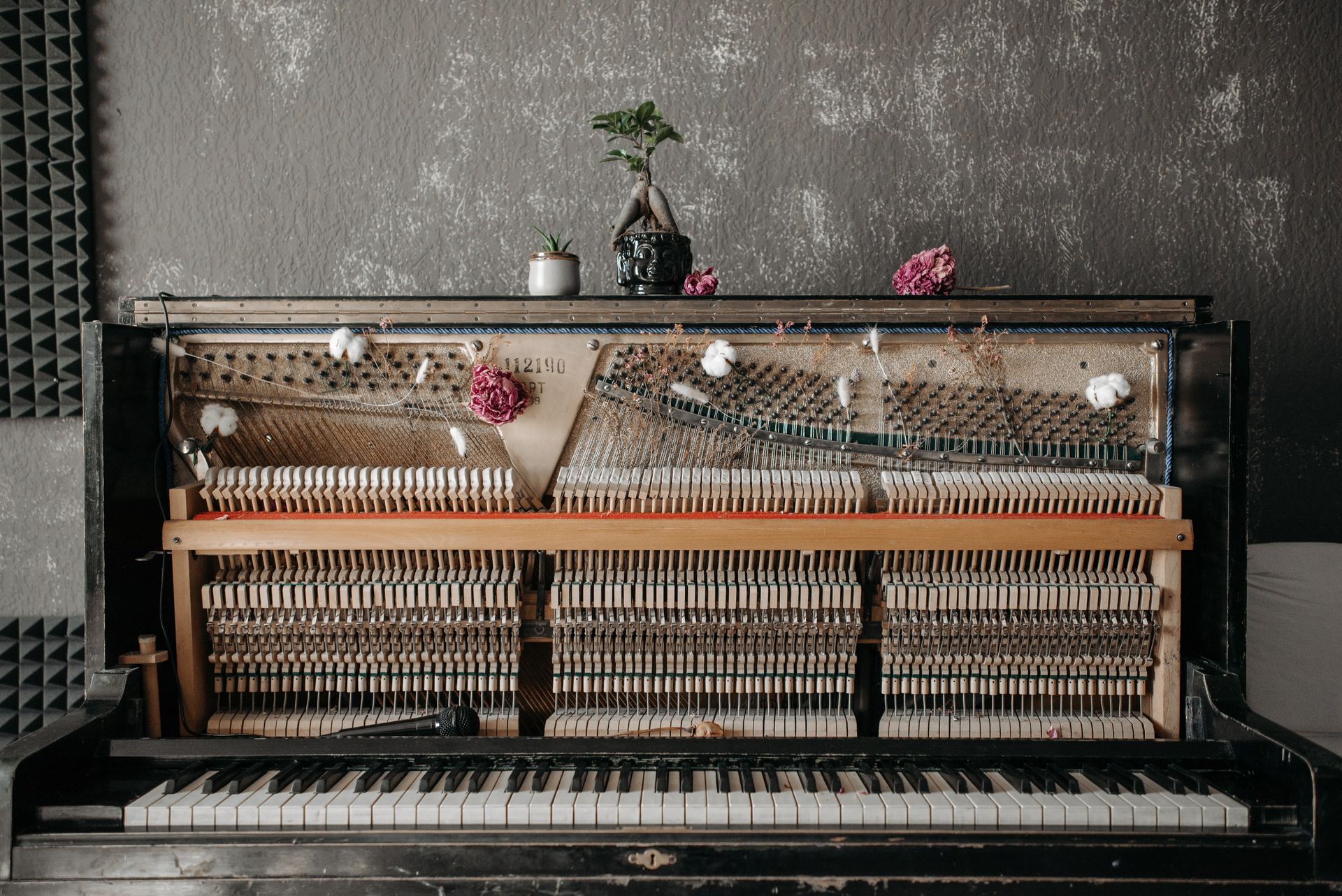 A vintage upright piano with exposed strings, adorned with flowers and a bonsai, in an acoustic studio setting.
