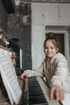 A smiling girl sits at a piano indoors, learning to play with music sheets.