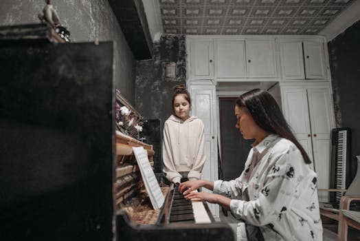 A student observes her teacher practicing piano in a cozy home music studio setting.