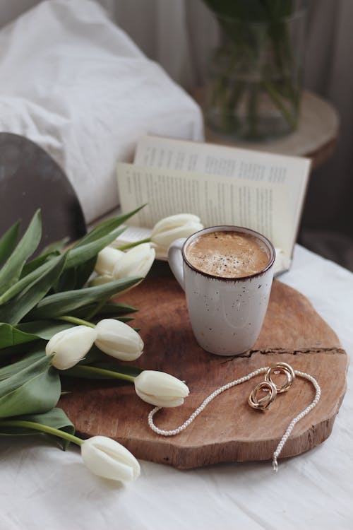 From above mug of fresh aromatic foamy chocolate with elegant jewelry and bunch of white tulips places on wooden tray on bed in morning
