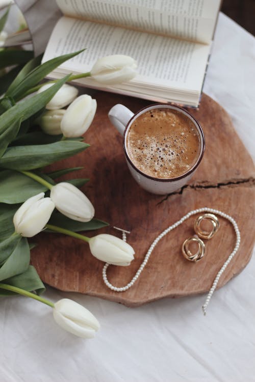 Free High angle of cup of hot chocolate with bouquet of fresh white tulips and stylish jewelry placed on wooden tray on bed near opened book at home Stock Photo